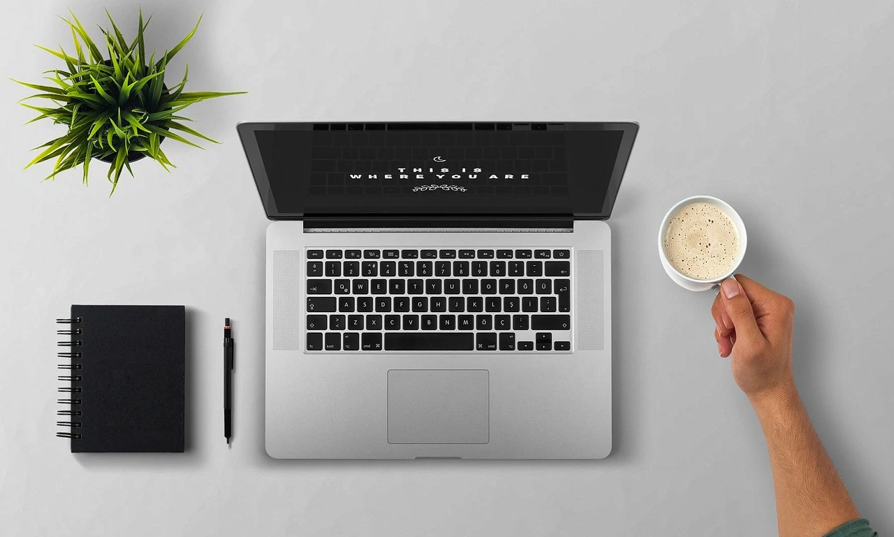 A desk seen from above with an open laptop, a notebook, a pen, a plant and a hand holding a cup of coffee.